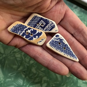 Könnte beinhalten: Drei Keramikknöpfe mit blau-weißem Blumenmuster. Die Knöpfe haben unterschiedliche Formen: rechteckig, trapezförmig und dreieckig. Jeder Knopf hat zwei Löcher zum Annähen.