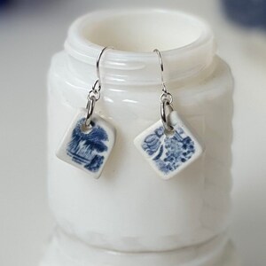 May include: A pair of dangle earrings featuring square-shaped white ceramic tiles with blue transferware-style landscape and floral patterns. The tiles are suspended from silver-toned metal ear wires by small circular jump rings.