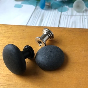 May include: Two black round cabinet knobs with a silver metal base. The knobs have a textured surface.