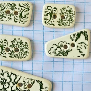 May include: Five off-white, rectangular buttons with dark green floral designs. Each button has two holes for sewing. The buttons are arranged on a blue grid background, showcasing their decorative patterns.