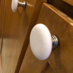 May include: Two white ceramic cabinet knobs with silver hardware mounted on a wooden cabinet door.