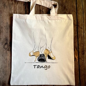 以下が含まれることがあります： 黒と金の絵の具で描かれたタンゴシューズを履いた2つの足の画像が描かれた白いキャンバス地のトートバッグ。靴の下に「タンゴ」という文字が金色の絵の具で書かれている。