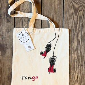 May include: A white canvas tote bag with black and red high heels printed on it. The bag has a black handle and a tag that says "Hand-made". The text "Tango" is printed in red at the bottom of the bag.