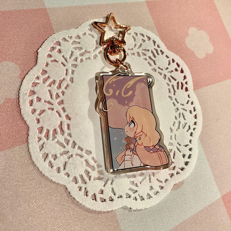 May include: A clear acrylic keychain featuring a cartoon illustration of a person with blonde hair looking at a cloudy sky. The keychain has a gold star-shaped clasp and is on a white lace doily.