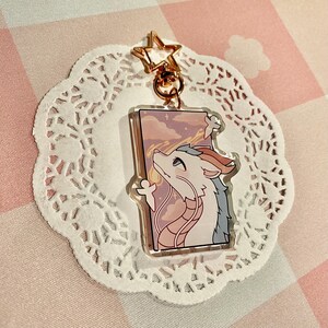 May include: A clear acrylic keychain featuring a cartoon dragon with white, blue, and peach tones, gazing upwards at a pink and purple sky. The keychain has a gold star-shaped clasp and a gold ring. The dragon is set against a rectangular background.