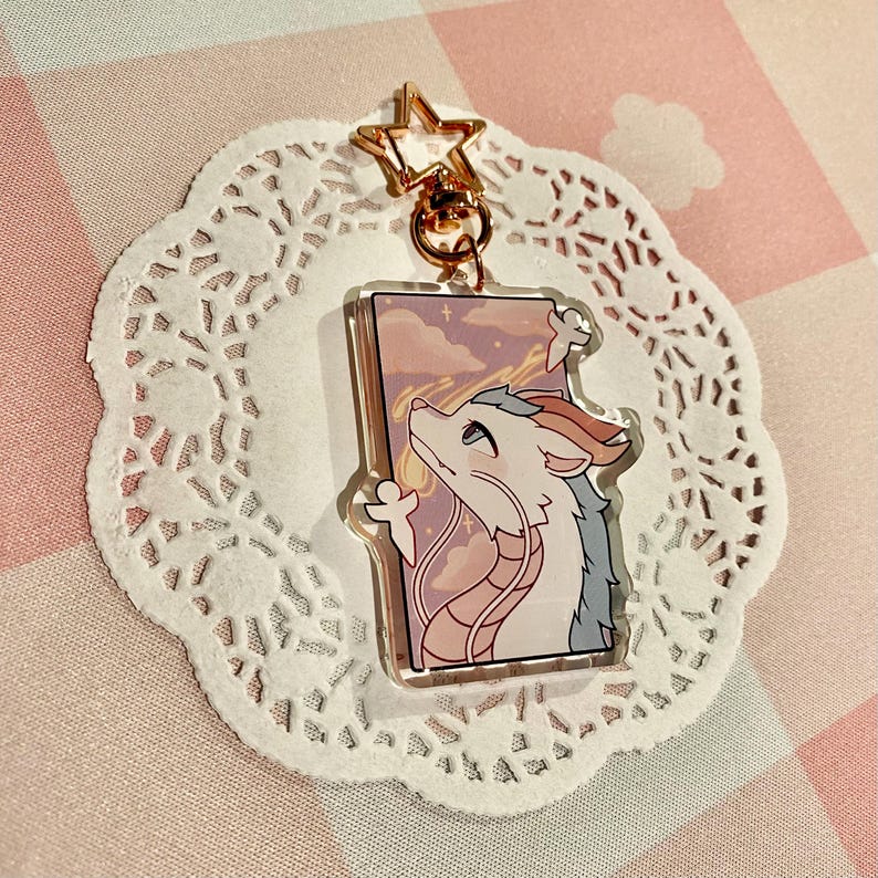 May include: A clear acrylic keychain featuring a cartoon dragon with white, blue, and peach tones, gazing upwards at a pink and purple sky. The keychain has a gold star-shaped clasp and a gold ring. The dragon is set against a rectangular background.