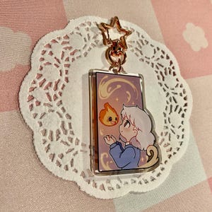 May include: A rectangular acrylic keychain featuring a cartoon illustration of a character with white hair and a blue outfit, interacting with a small, orange flame. The keychain has a gold star-shaped clasp and is set against a white lace doily.