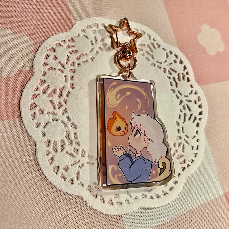 May include: A rectangular acrylic keychain featuring a cartoon illustration of a character with white hair and a blue outfit, interacting with a small, orange flame. The keychain has a gold star-shaped clasp and is set against a white lace doily.