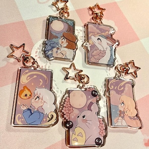 May include: A collection of acrylic keychains featuring anime-style illustrations. Each keychain has a gold-colored star-shaped clasp. The designs include characters and scenes with soft color palettes, such as a moonlit scene and a dragon.
