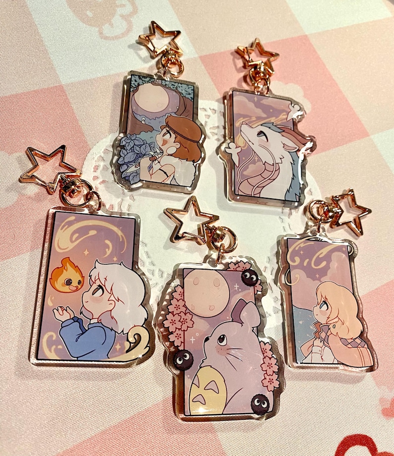 May include: A collection of acrylic keychains featuring anime-style illustrations. Each keychain has a gold-colored star-shaped clasp. The designs include characters and scenes with soft color palettes, such as a moonlit scene and a dragon.