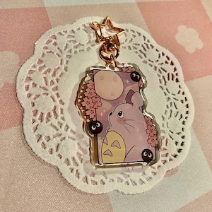 May include: A clear acrylic charm featuring a lavender-colored cartoon character with a full moon and cherry blossoms. The charm has a gold-colored star-shaped clasp and a small chain. The background is a pink and white checkered pattern.