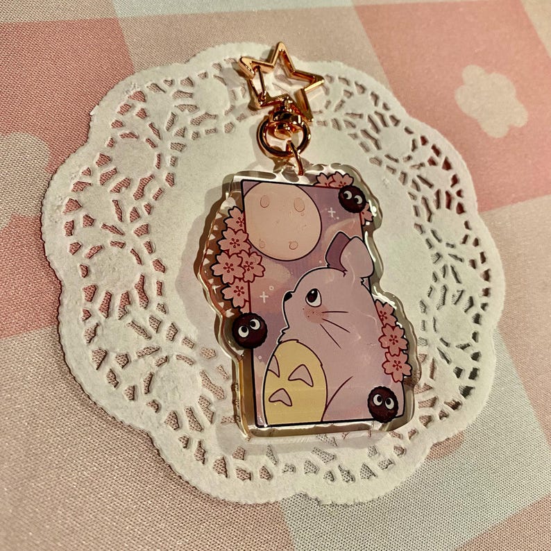 May include: A clear acrylic charm featuring a lavender-colored cartoon character with a full moon and cherry blossoms. The charm has a gold-colored star-shaped clasp and a small chain. The background is a pink and white checkered pattern.