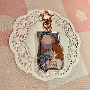 May include: A clear acrylic keychain with a cartoon illustration of a person looking at a moonlit scene. The keychain has a gold star-shaped clasp and a gold ring. The background is a pink and white checkered pattern.