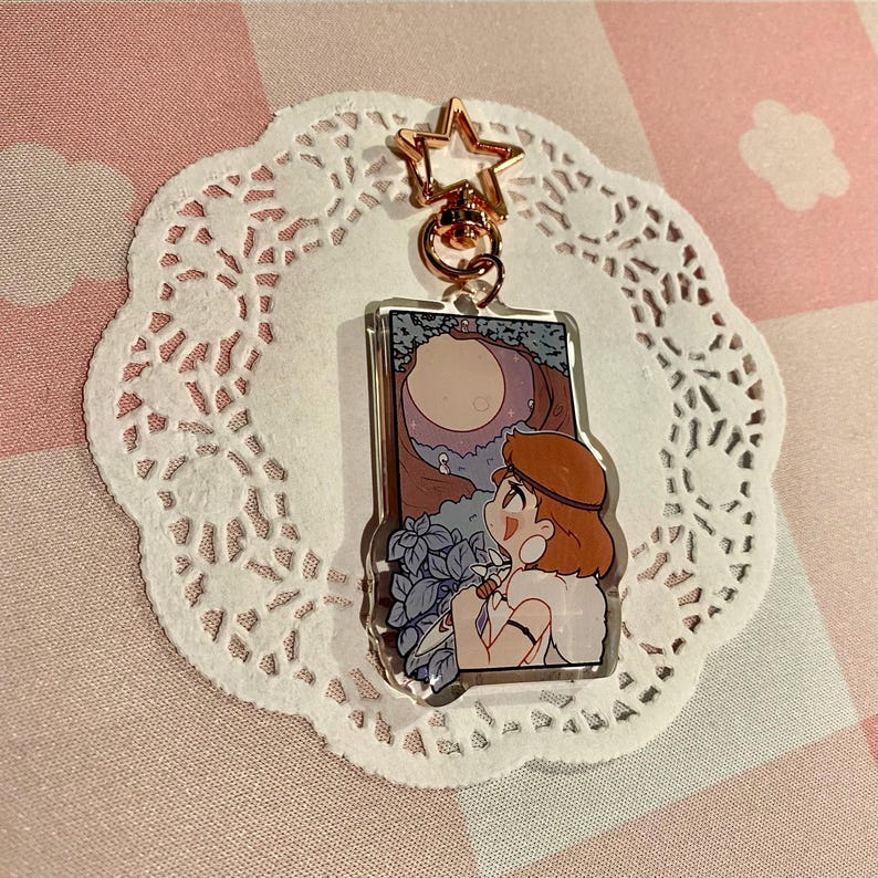 May include: A clear acrylic keychain with a cartoon illustration of a person looking at a moonlit scene. The keychain has a gold star-shaped clasp and a gold ring. The background is a pink and white checkered pattern.