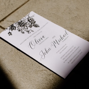 May include: A white wedding program with black floral illustrations and elegant script. The program lists the names Olivia and John Michael, along with the date, time, and location of the ceremony: St. Mary's Catholic Church, College Station, Texas.