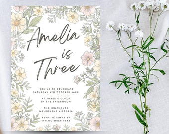 May include: A birthday invitation with the text "Amelia is Three." The invitation is decorated with pastel-colored flowers and greenery. The invitation includes celebration details. A sprig of white flowers is placed to the right of the invitation.