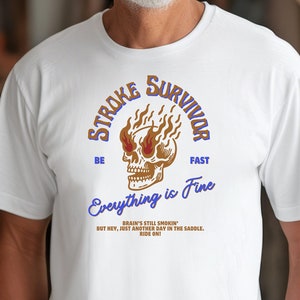May include: White t-shirt with a graphic of a skull with flames and the text "Stroke Survivor" above it and "Everything is Fine" below it. The text "Be Fast" is on either side of the skull. The text below the skull reads "Brain's still smorkin' but hey, just another day in the saddle. Ride on!"