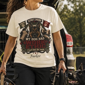 May include: A cream-coloured t-shirt featuring a graphic of a German Shepherd, the text "SCHUTZHUND", "MY DOG HAS WORK ETHIC", "I just hold the leash", and the name "Parker". The design includes paw prints, a dumbbell, and a tracking flag.