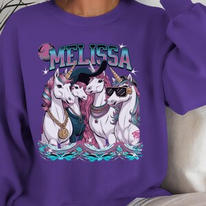 May include: Purple sweatshirt with a graphic of three unicorns wearing sunglasses, hats, and jewelry. The text "MELISSA" is printed above the unicorns.