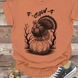 May include: Orange t-shirt with a black graphic of a turkey perched on a pumpkin, with the text "F-CAW-F". The shirt is paired with gray sneakers with white laces. A bouquet of flowers is in the lower left corner.
