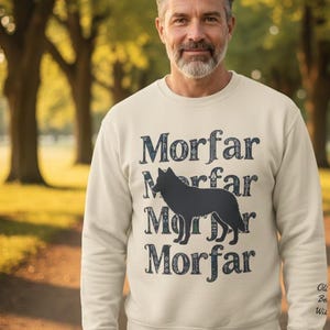 May include: A cream-colored sweatshirt with the word "Morfar" in a dark blue, vintage font, repeated four times with a black dog silhouette in the center. The sweatshirt is worn by a person in a park setting.
