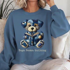 May include: A blue sweatshirt featuring a dog illustration made of patchwork denim, reading a book. The text "Dogs. Books. Quilting" is printed below the dog. The sweatshirt has long sleeves and a crew neck.