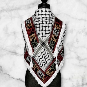 Puede incluir: Un pañuelo keffiyeh blanco, negro y rojo con patrones geométricos y flecos. El pañuelo se muestra sobre una cabeza y hombros de maniquí negro. El diseño incluye escritura árabe y un patrón de cuadros.