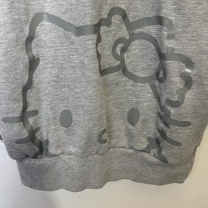 May include: A gray sweatshirt featuring a large, light gray outline of a cartoon cat's face. The design includes the cat's eyes, nose, whiskers, and bow. The sweatshirt has a ribbed hem and cuffs.