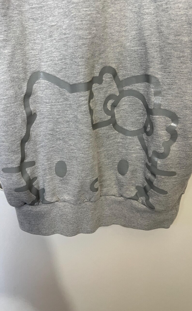May include: A gray sweatshirt featuring a large, light gray outline of a cartoon cat's face. The design includes the cat's eyes, nose, whiskers, and bow. The sweatshirt has a ribbed hem and cuffs.