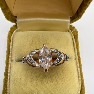 Puede incluir: Un anillo de oro con una gran piedra preciosa transparente de talla marquesa como pieza central. El anillo tiene piedras más pequeñas y transparentes y detalles azules a ambos lados de la piedra principal, en un joyero forrado de terciopelo.