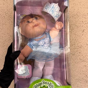 Peut inclure: Poupée Cabbage Patch Kids Ice Skating Star dans une boîte en plastique transparent. La poupée porte une robe bleue avec des motifs floraux blancs et une couronne assortie. La boîte a un logo vert et blanc.