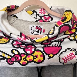 May include: A gray hoodie with a Hello Kitty design. The white cowl neck features a repeating pattern of pink, yellow, and black Hello Kitty graphics. The Hello Kitty logo is on a tag at the neckline.