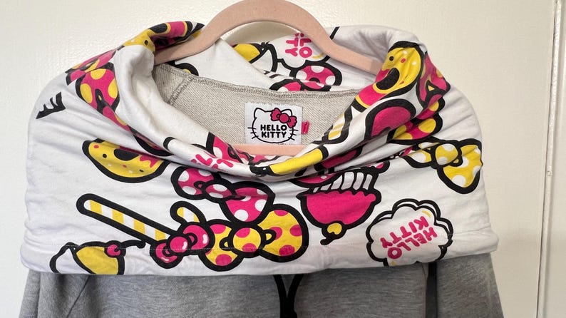 May include: A gray hoodie with a Hello Kitty design. The white cowl neck features a repeating pattern of pink, yellow, and black Hello Kitty graphics. The Hello Kitty logo is on a tag at the neckline.