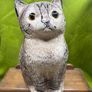 May include: A vintage cat figurine with a distressed, textured finish. The cat is primarily cream-colored with dark brown stripes and large, amber-colored eyes. It is sitting on a small wooden cabinet.