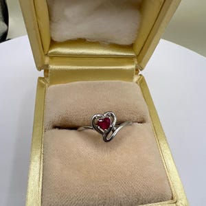 May include: A silver-toned ring featuring a heart-shaped red gemstone set within a swirling metal band. The ring is displayed inside a rectangular jewelry box with a light yellow exterior and a soft, beige velvet-textured interior lining.