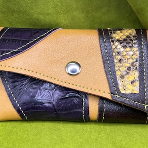 Può includere: Un portafoglio rettangolare in pelle con base color cuoio e patta diagonale. Il design presenta un pannello viola scuro con texture coccodrillo, una striscia verticale in pelle con motivo a pelle di serpente gialla e nera e un bottone a pressione argentato.