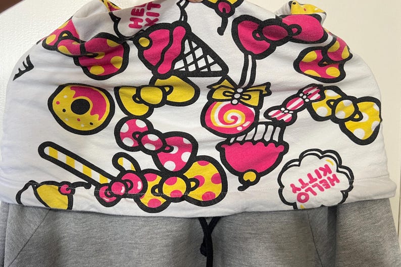 May include: A gray hoodie with a white hood featuring a colorful pattern of donuts, bows, ice cream cones, and candy. The hood design includes the text "HELLO KITTY" in a cloud-shaped design.