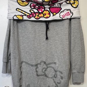 May include: Gray sweatshirt dress featuring a Hello Kitty graphic. The top has a white cowl neck with a colorful pattern of pink, yellow, and red bows and candies. The Hello Kitty logo is visible on the cowl neck.