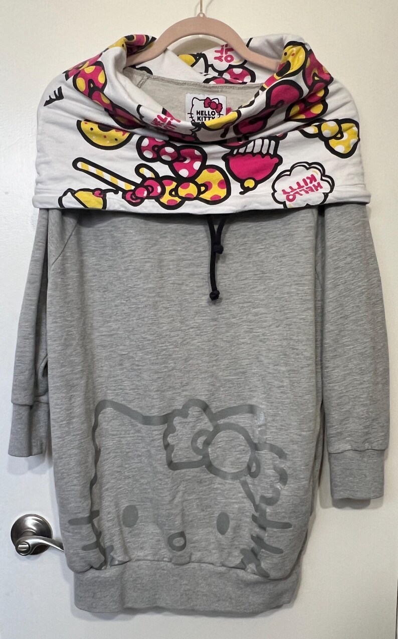 May include: Gray sweatshirt dress featuring a Hello Kitty graphic. The top has a white cowl neck with a colorful pattern of pink, yellow, and red bows and candies. The Hello Kitty logo is visible on the cowl neck.