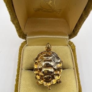 May include: A gold-colored turtle pendant inside a vintage velvet jewelry box. The turtle has a detailed shell and small legs. The box is a mustard yellow color with a satin lining and a crest on the inside of the lid.