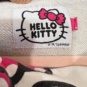 May include: Close-up of a gray sweatshirt label featuring the Hello Kitty logo with a pink bow. The label also includes the text "HELLO KITTY" and the copyright information.