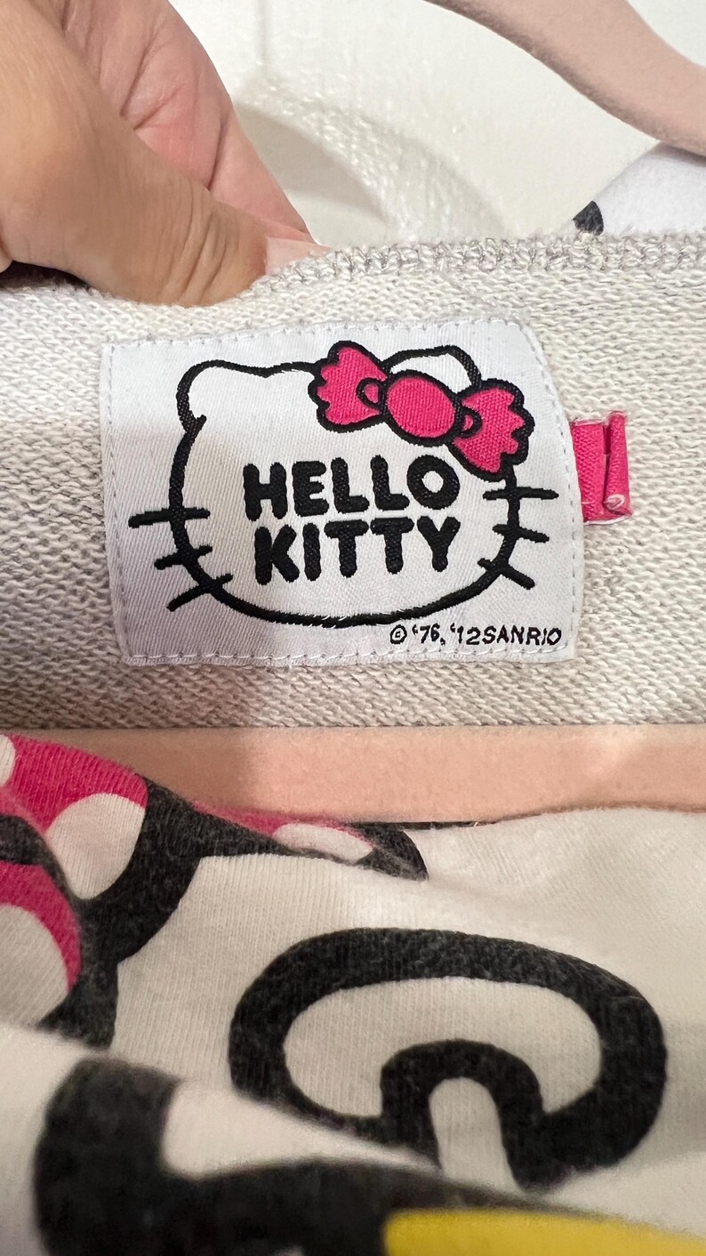 May include: Close-up of a gray sweatshirt label featuring the Hello Kitty logo with a pink bow. The label also includes the text "HELLO KITTY" and the copyright information.