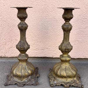 Puede incluir: Un par de candelabros antiguos de latón con diseños ornamentados. Cada candelabro tiene una base ancha, un tallo decorado y una parte superior acampanada. El latón tiene una pátina cálida y envejecida. Están sobre una superficie gris.