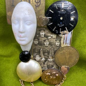 Könnte beinhalten: Eine Mixed-Media-Assemblage-Brosche mit einem weißen Harzgesicht, einem schwarzen Technos Sky Light-Zifferblatt, einem alten Foto von Soldaten, einem Messingschild mit der Nummer 1119, einer Messingmarke und Goldketten.