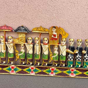 Puede incluir: Una escena de procesión balinesa de madera tallada. Las figuras visten trajes tradicionales, portando paraguas, un palanquín e instrumentos musicales. La base está decorada con un colorido patrón geométrico.