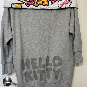 May include: Gray sweatshirt dress featuring the text "HELLO KITTY" in a darker gray. The collar is white with a colorful pattern of donuts, bows, and the words "HELLO KITTY".