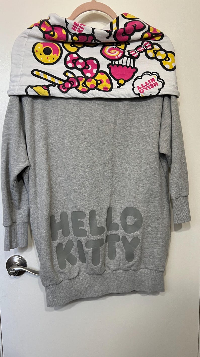 May include: Gray sweatshirt dress featuring the text "HELLO KITTY" in a darker gray. The collar is white with a colorful pattern of donuts, bows, and the words "HELLO KITTY".