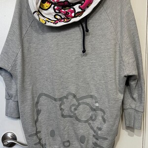 May include: Gray sweatshirt with a Hello Kitty graphic on the front. The cowl neck features a white, pink, and yellow pattern with the words "Hello Kitty" printed on it. The sweatshirt has three-quarter sleeves.