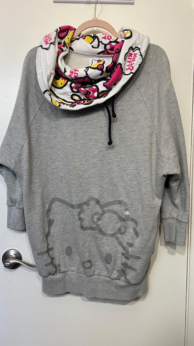May include: Gray sweatshirt with a Hello Kitty graphic on the front. The cowl neck features a white, pink, and yellow pattern with the words "Hello Kitty" printed on it. The sweatshirt has three-quarter sleeves.