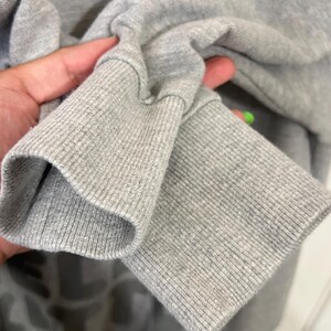 May include: Close-up of a gray sweatshirt sleeve. The ribbed cuff is visible, along with the soft, textured fabric of the sleeve. The sweatshirt appears to be made of a comfortable material, suitable for casual wear.